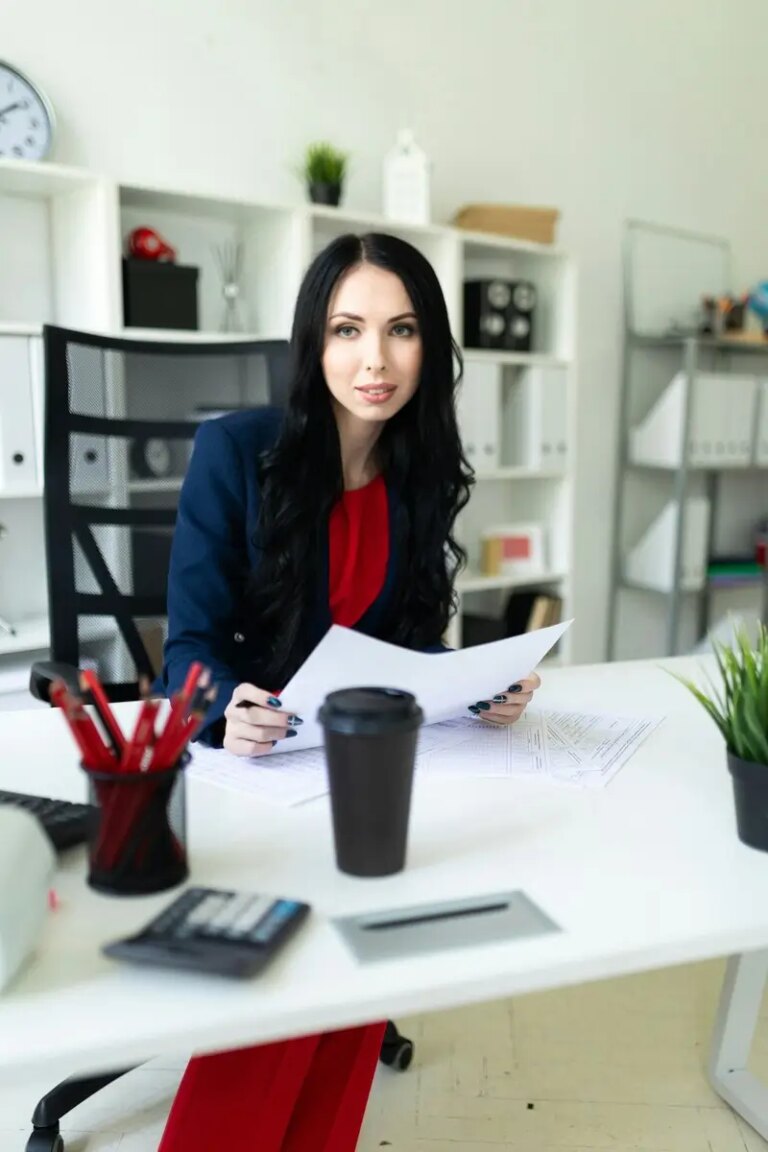 Beautiful young girl is looking through documents, sitting in the office at the table.