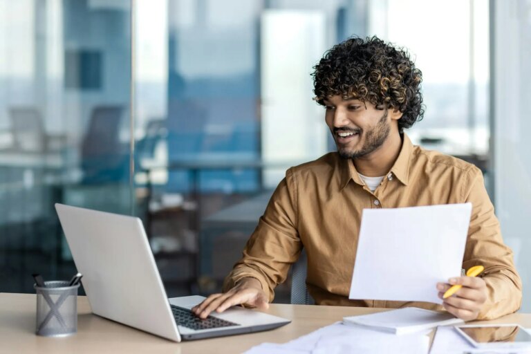 Smiling arab employee using personal computer while holding document and pen in hand at workspace